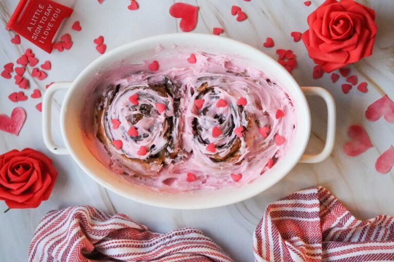 Valentine cinnamon rolls with pink frosting and heart sprinkles partially visible beneath overlay graphics.