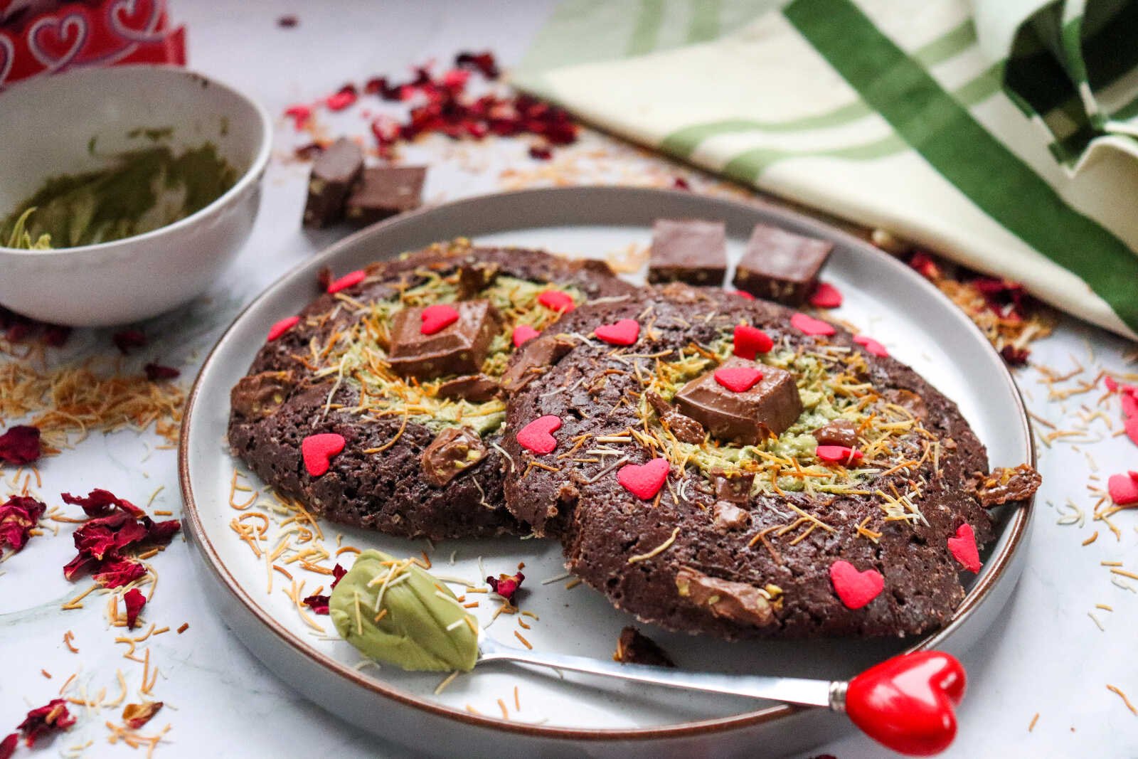 Dubai chocolate cookies topped with pistachio cream, kataifi strands, chocolate chunks, and red heart sprinkles on a plate.